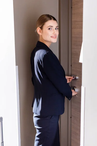 Joyful businesswoman in formal wear holding room card while unlocking door in hotel — Stock Photo