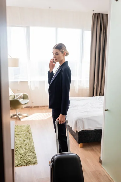 Businesswoman in suit standing with luggage and talking on smartphone in hotel room — Stock Photo