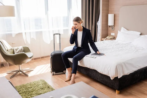 Selective focus of businesswoman in suit talking on smartphone while sitting on bed near luggage in hotel — Stock Photo