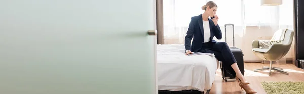 Horizontales Bild einer Geschäftsfrau im Anzug, die auf dem Smartphone spricht, während sie im Hotelbett neben dem Gepäck sitzt — Stockfoto
