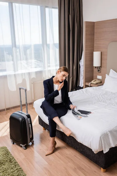 Молода бізнес-леді в костюмі говорить по смартфону, сидячи на ліжку біля багажу в готелі — стокове фото