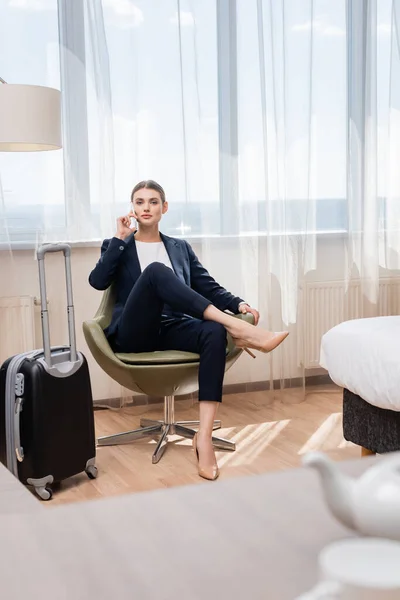 Joven mujer de negocios en traje hablando en el teléfono inteligente mientras está sentado en el sillón cerca de equipaje en el hotel - foto de stock