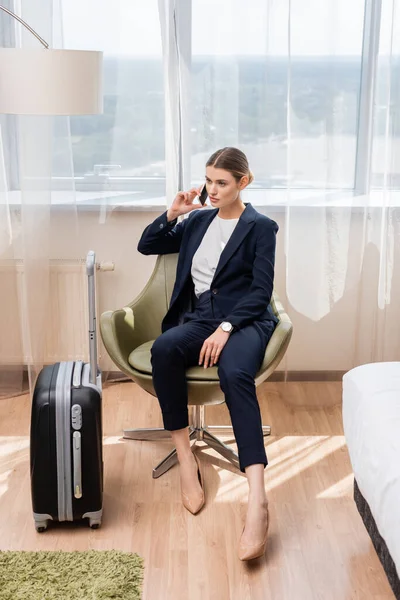 Joven mujer de negocios en traje hablando en teléfono inteligente y sentado en sillón cerca de equipaje en el hotel - foto de stock