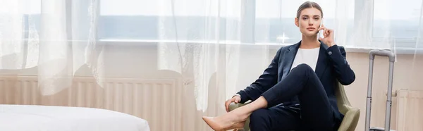 Raccolto panoramico di donna d'affari in giacca e cravatta parlare su smartphone e seduto in poltrona vicino ai bagagli in hotel — Foto stock