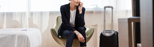 Horizontal crop of businesswoman in suit talking on smartphone and sitting in armchair near baggage in hotel — Stock Photo