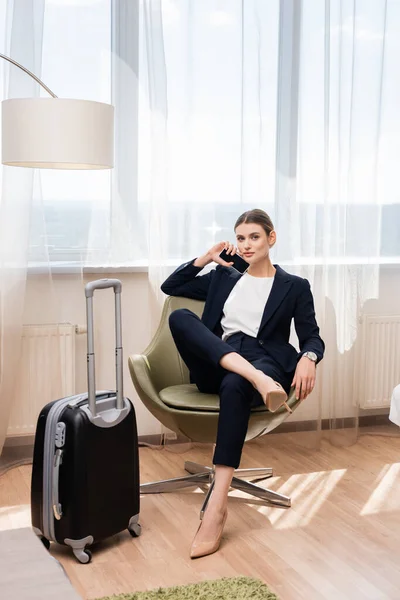 Joven mujer de negocios en traje sosteniendo teléfono inteligente con pantalla en blanco y sentado en sillón cerca de equipaje en el hotel - foto de stock