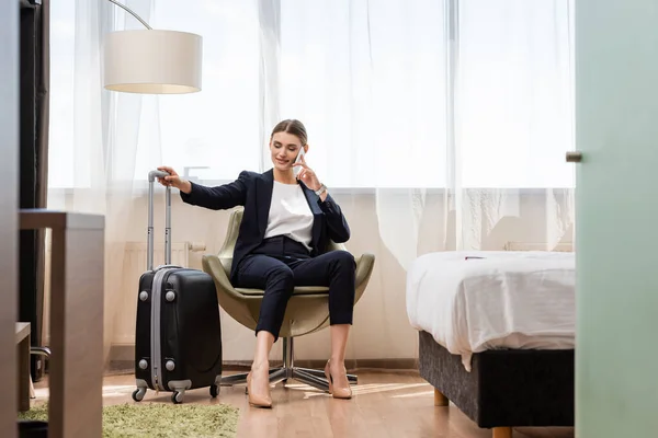 Enfoque selectivo de la mujer de negocios complacido en traje hablando en el teléfono inteligente y sentado en sillón cerca de equipaje en el hotel - foto de stock