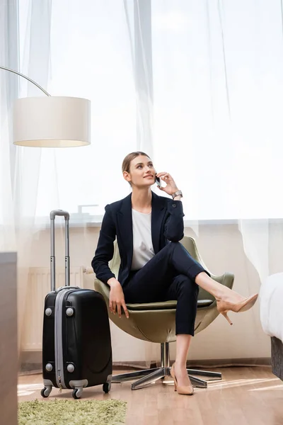 Joven mujer de negocios en traje hablando en teléfono inteligente y sentado en sillón cerca de equipaje en el hotel - foto de stock