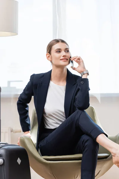 Alegre mujer de negocios en traje hablando en el teléfono inteligente y sentado en sillón cerca de la bolsa de viaje en el hotel - foto de stock