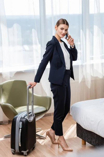 Empresária de terno de pé com bagagem enquanto conversa no smartphone no quarto de hotel — Fotografia de Stock