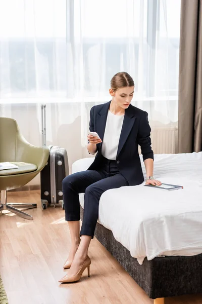 Businesswoman in suit holding smartphone and looking at notebook with air ticket near baggage in hotel room — Stock Photo