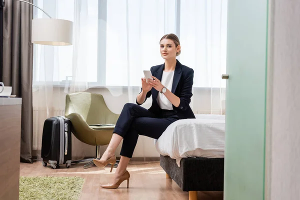 Молода бізнес-леді в костюмі тримає смартфон, сидячи на ліжку акуратна дорожня сумка в готельному номері — стокове фото