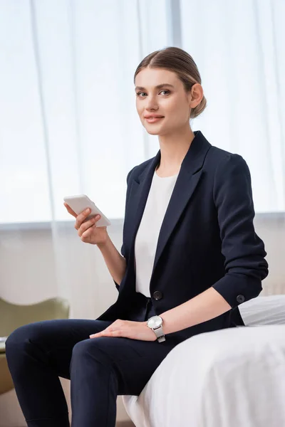 Geschäftsfrau im Anzug hält Handy in der Hand und blickt in die Kamera, während sie im Hotelzimmer auf dem Bett sitzt — Stockfoto