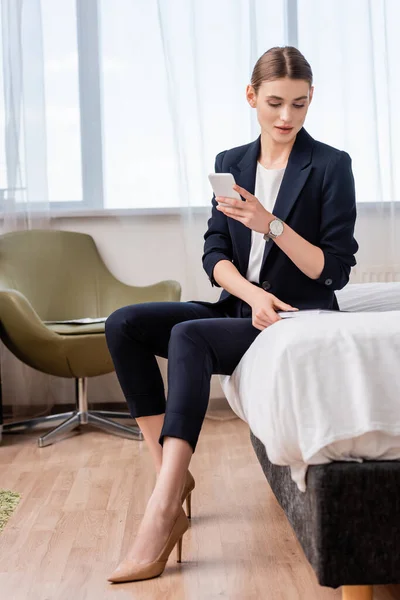 Бізнес-леді в офіційному одязі тримає смартфон і дивиться на блокнот в готельному номері — стокове фото