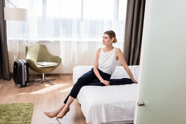 Mujer de negocios mirando hacia otro lado mientras está sentada en la cama en la habitación de hotel moderna - foto de stock