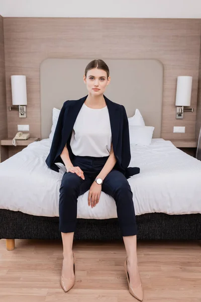 Young businesswoman in suit looking at camera and sitting on bed in hotel — Stock Photo