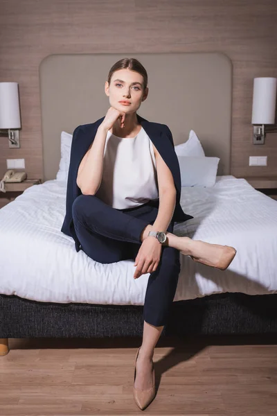 Femme d'affaires en costume regardant la caméra et assis sur le lit dans l'hôtel — Photo de stock
