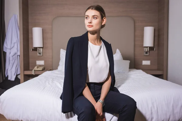 Joven mujer de negocios en traje mirando hacia otro lado y sentada en la cama en el hotel - foto de stock
