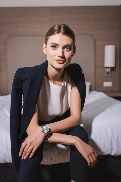 Joven mujer de negocios en ropa formal mirando a la cámara y sentado en la cama en la habitación de hotel - foto de stock