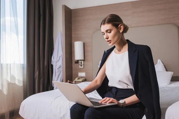 Женщина в костюме сидит на кровати и смотрит на ноутбук в номере отеля — стоковое фото