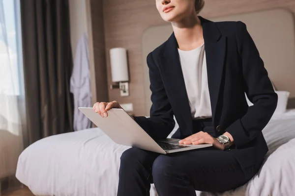Обрезанный вид женщины в костюме, сидящей на кровати с ноутбуком в номере отеля — стоковое фото