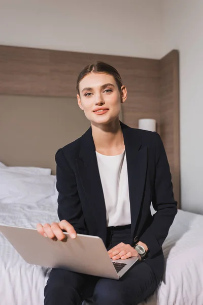 Joyful woman in suit sitting on bed with laptop and looking at camera in hotel room — Stock Photo