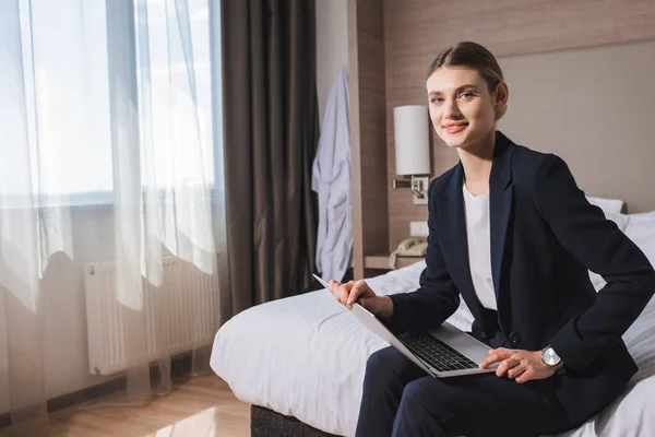 Pleased young woman in suit sitting on bed with laptop in hotel room — Stock Photo