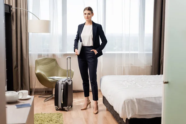 Selective focus of young businesswoman standing with hand in pocket and holding laptop near travel bag in hotel room — Stock Photo