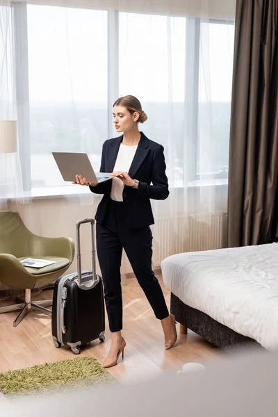 Молодая деловая женщина, стоящая и использующая ноутбук возле дорожной сумки в номере отеля — стоковое фото