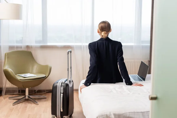 Vue arrière de la femme d'affaires assise sur le lit près d'un ordinateur portable avec écran vierge et valise dans la chambre d'hôtel — Photo de stock