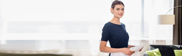 Récolte panoramique de femme de chambre joyeuse tenant serviette propre et regardant la caméra dans la chambre d'hôtel — Photo de stock