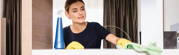 Panoramic crop of maid in rubber gloves holding rag while cleaning shelf in hotel room — Stock Photo