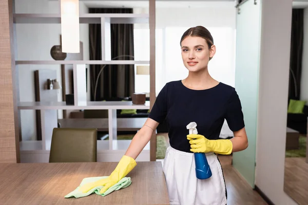 Ama de llaves en delantal y guantes de goma que sostienen la botella y el trapo mientras limpian la superficie en la habitación del hotel — Stock Photo