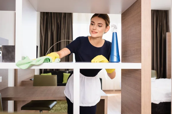 Maid in apron and rubber gloves rag while cleaning shelf in hotel room — Stock Photo