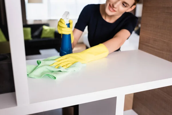 Enfoque selectivo de la criada en guantes de goma sosteniendo la botella de aerosol y trapo mientras se limpia el estante en la habitación de hotel - foto de stock
