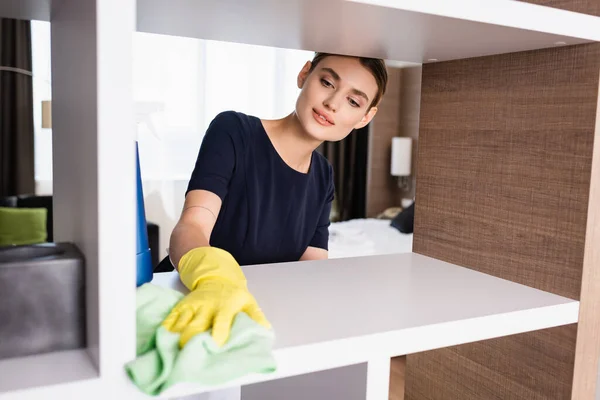 Selective focus of maid in rubber gloves holding spray bottle and rag while cleaning shelf in hotel room — Stock Photo