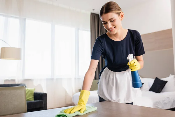 Chambermaid in apron and rubber gloves holding spray bottle and rag while cleaning wooden surface in hotel room — Stock Photo