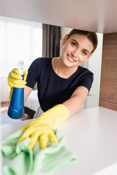 Enfoque selectivo de la criada alegre en guantes de goma que sostienen la botella y el trapo mientras limpian el estante en la habitación del hotel - foto de stock