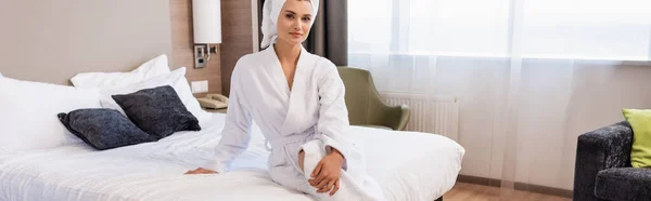 Горизонтальний урожай жінки в білому рушнику і халаті, що сидить на ліжку в готельному номері — стокове фото