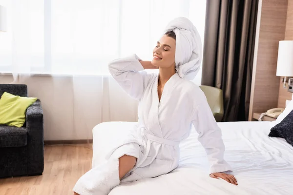 Mujer alegre con los ojos cerrados en toalla blanca y albornoz sentado en la cama en la habitación del hotel - foto de stock