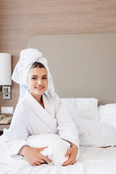 Mulher contente em toalha e roupão de banho branco deitado na cama e olhando para a câmera no quarto do hotel — Fotografia de Stock