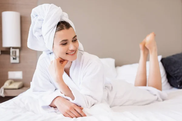 Descalça e mulher satisfeita em toalha e roupão de banho branco deitado na cama no quarto do hotel — Fotografia de Stock