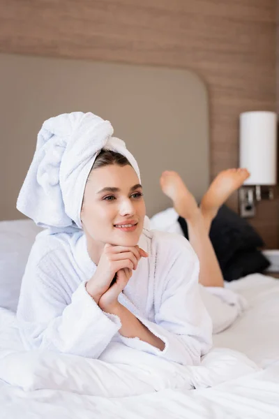 Mulher descalça em toalha e roupão de banho branco deitado na cama e olhando para longe no quarto do hotel — Fotografia de Stock