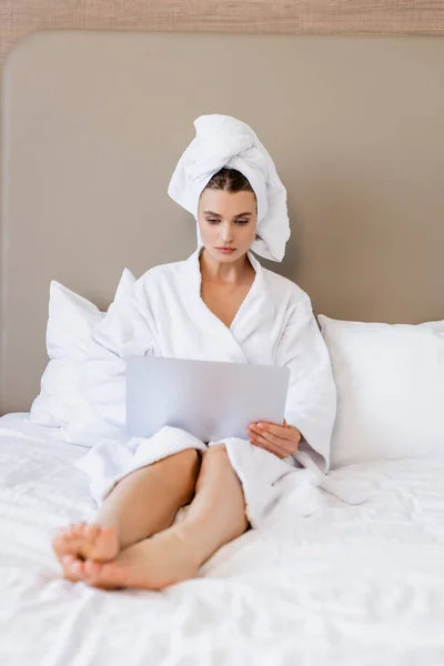 Selective focus of barefoot woman in bathrobe and towel using laptop on bed — Stock Photo