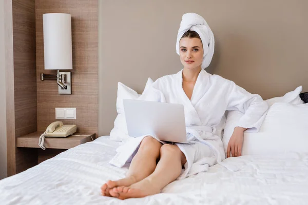 Selective focus of barefoot young woman in bathrobe using laptop on bed — Stock Photo