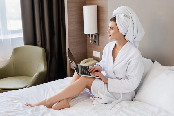 Barefoot young freelancer in bathrobe using laptop on bed — Stock Photo