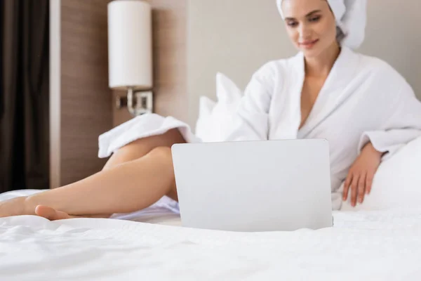 Selektiver Fokus der jungen Frau im Bademantel mit Laptop auf dem Bett — Stockfoto