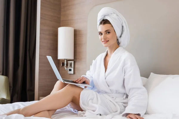 Young freelancer in bathrobe and towel using laptop on bed — Stock Photo