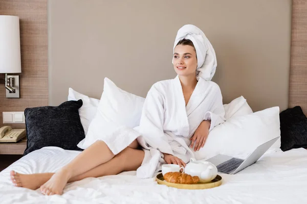 Молода жінка в халаті лежить біля ноутбука і підносу сніданку на ліжку — стокове фото