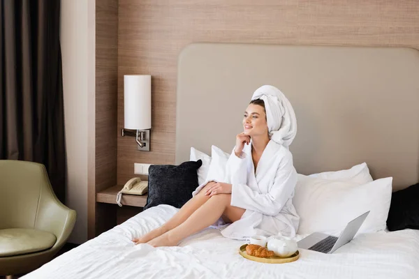 Junge Frau im Bademantel sitzt neben Laptop und Frühstückstablett auf dem Bett — Stockfoto